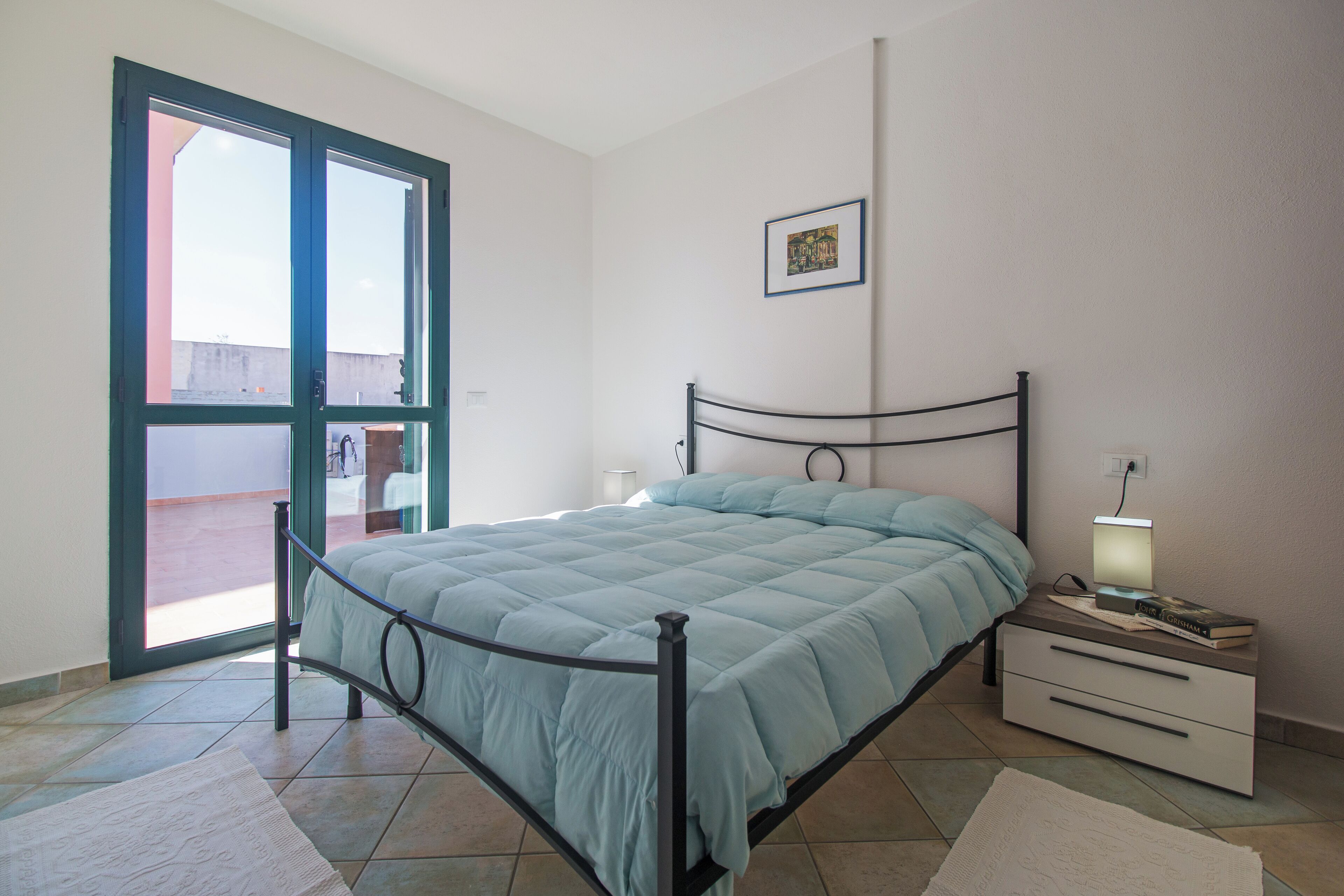 A bedroom with a bed, a bedside table with a lamp, a framed picture on the wall, and a sliding glass door leading to an outdoor area.