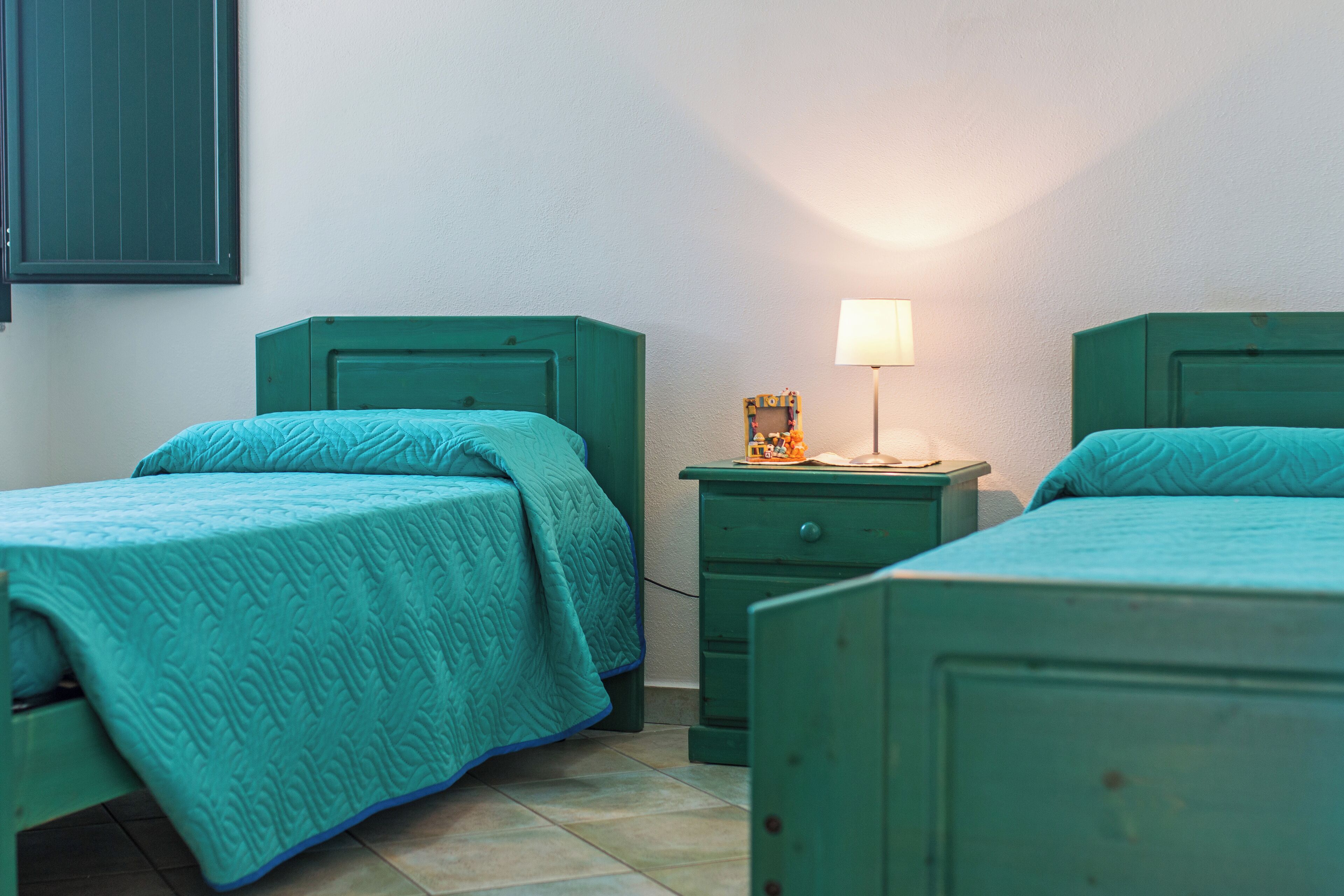 Una habitación con dos camas verdes, una mesita de noche con lámpara y una pequeña mesa con un marco de fotos.