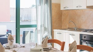 Un comedor con una mesa dispuesta para cuatro, una cocina con fregadero y horno, y vista a través de un ventanal.