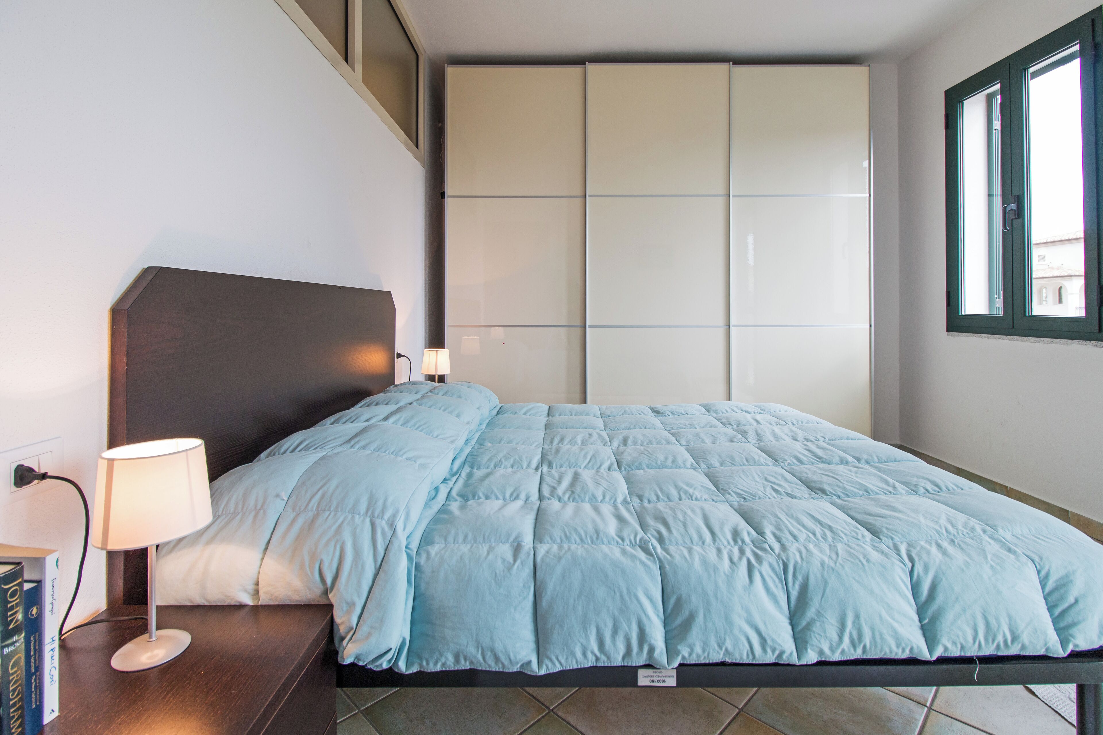 Een net opgemaakt bed met een lichtblauwe dekbedovertrek, een bedlampje en een nachtkastje met boeken.