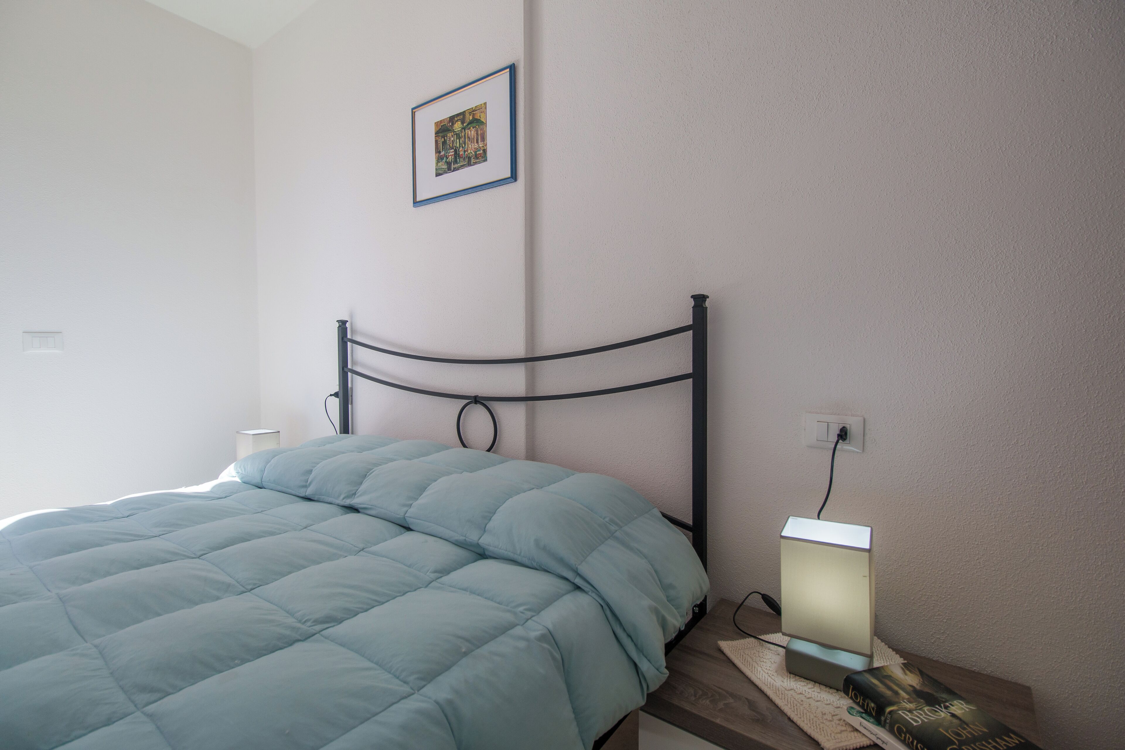 A neatly made bed with a blue comforter and a black metal headboard. A bedside table with a lamp and a book is visible.
