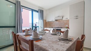 Un comedor con una mesa dispuesta para cuatro, sillas, una ventana con cortinas y una cocina al fondo.
