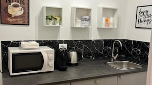 Une cuisine moderne avec un micro-ondes blanc, une machine à café et une bouilloire posés sur le plan de travail. Au-dessus du plan de travail, on trouve trois étagères blanches, chacune contenant différents objets.