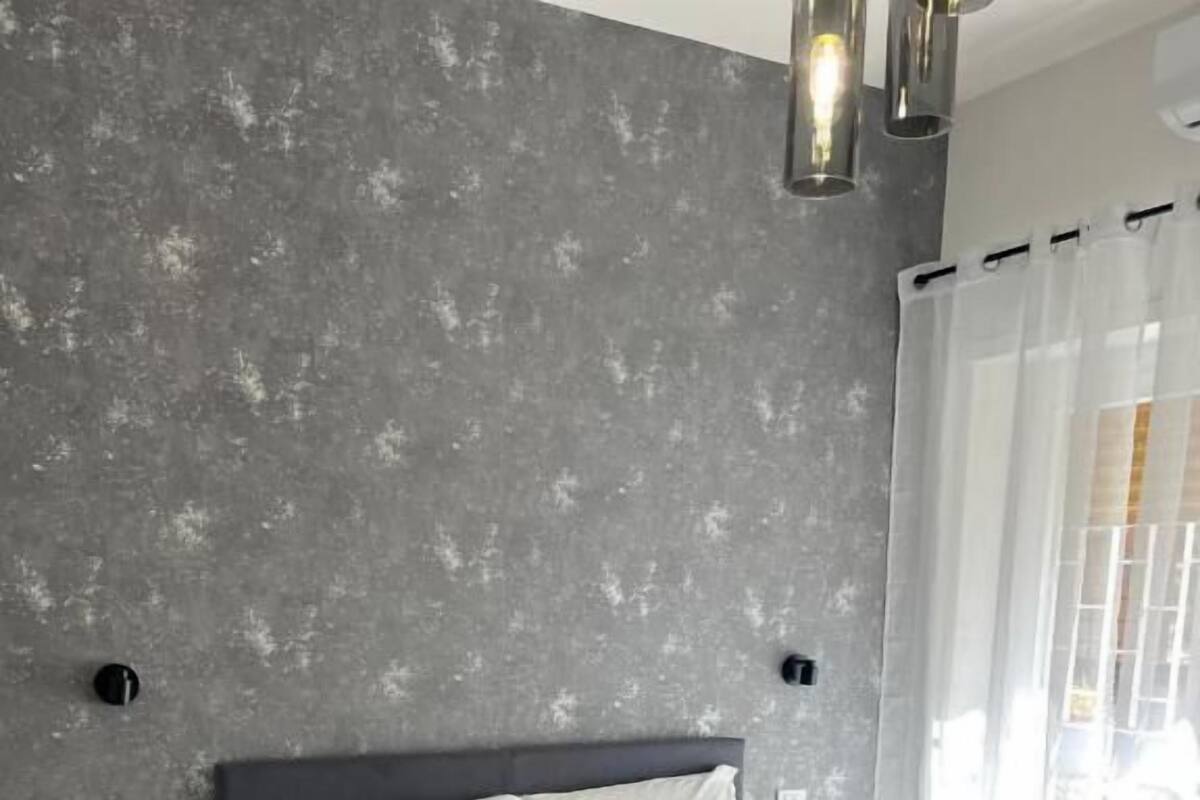 Un dormitorio moderno con pared gris texturizada, cabecero oscuro, ropa de cama blanca y una toalla blanca doblada con cuidado sobre la cama.