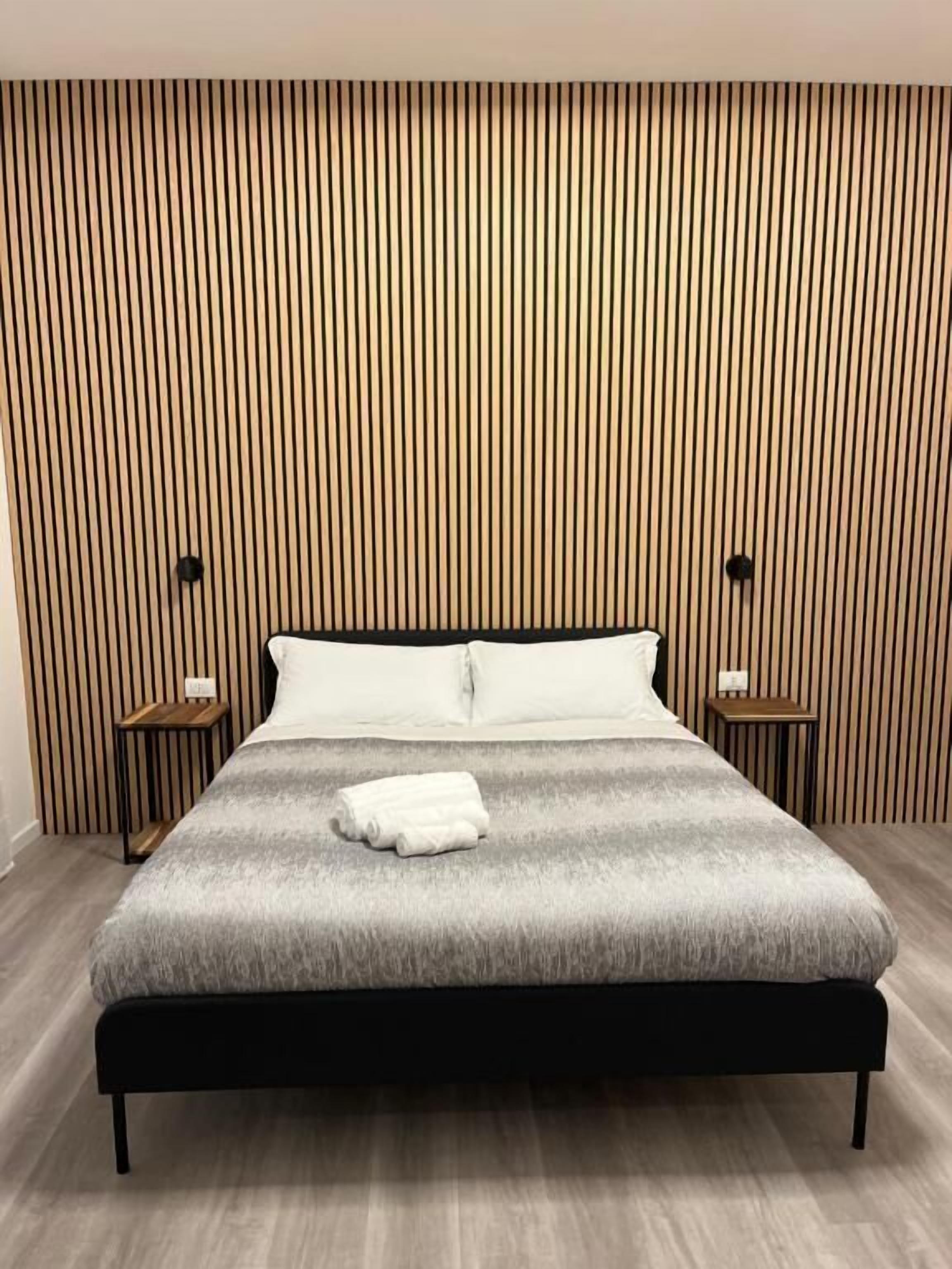 A modern bedroom with a large bed, wooden slat headboard, two bedside tables, and a folded towel on the bed.