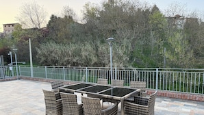 Un patio al aire libre con una mesa de vidrio y sillas de mimbre, rodeado por una cerca de metal y vistas a árboles y edificios.