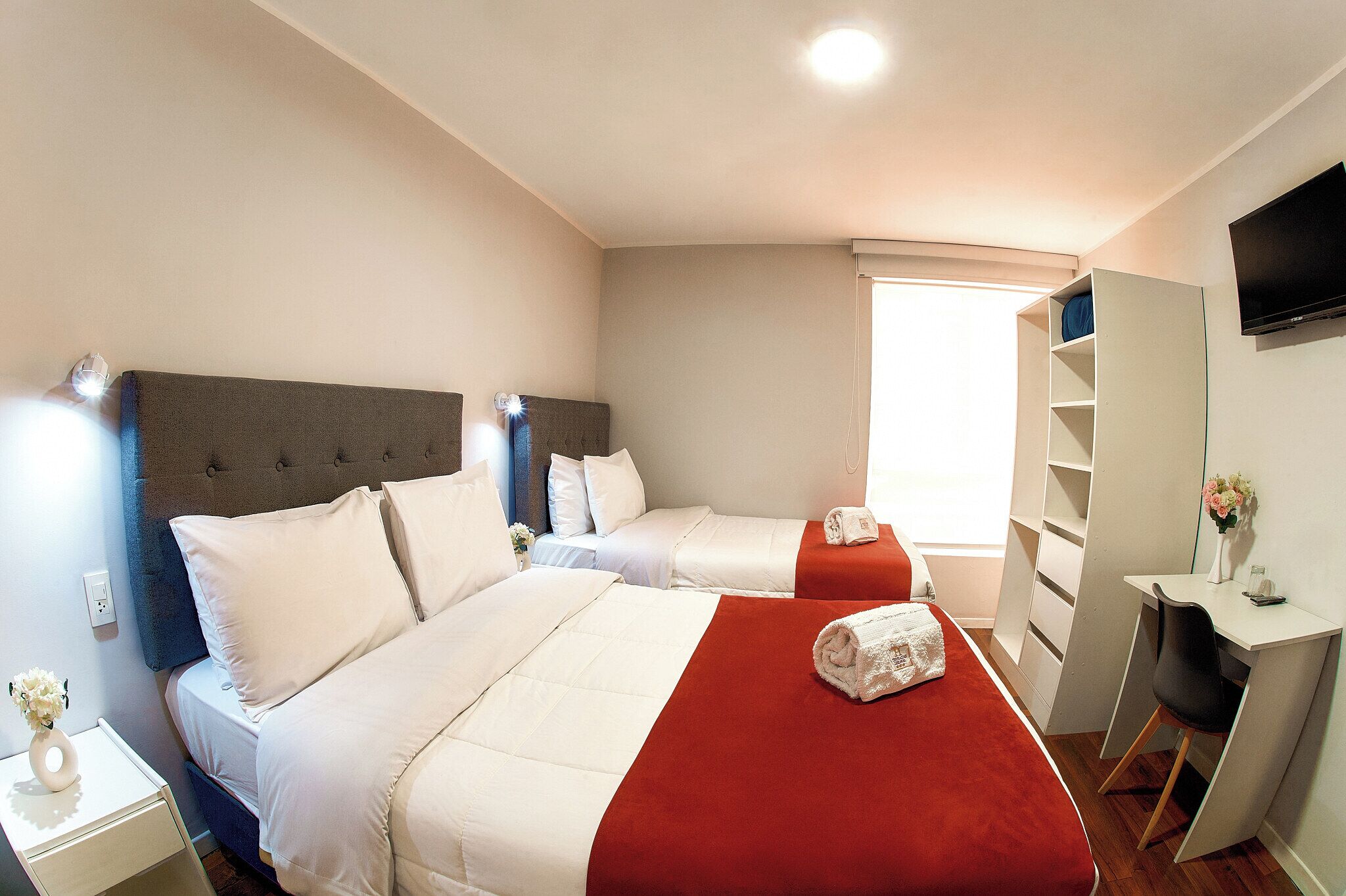 A hotel room with two beds, a desk, a TV, and a window.