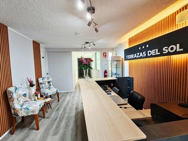 A reception area with a long wooden reception desk, two floral chairs, a black office chair, and a wall with vertical wooden slats and a sign that reads "TERRAZAS DEL SOL".