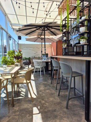 An outdoor café with tables and chairs, a bar area with shelves of plants, and a large umbrella providing shade.