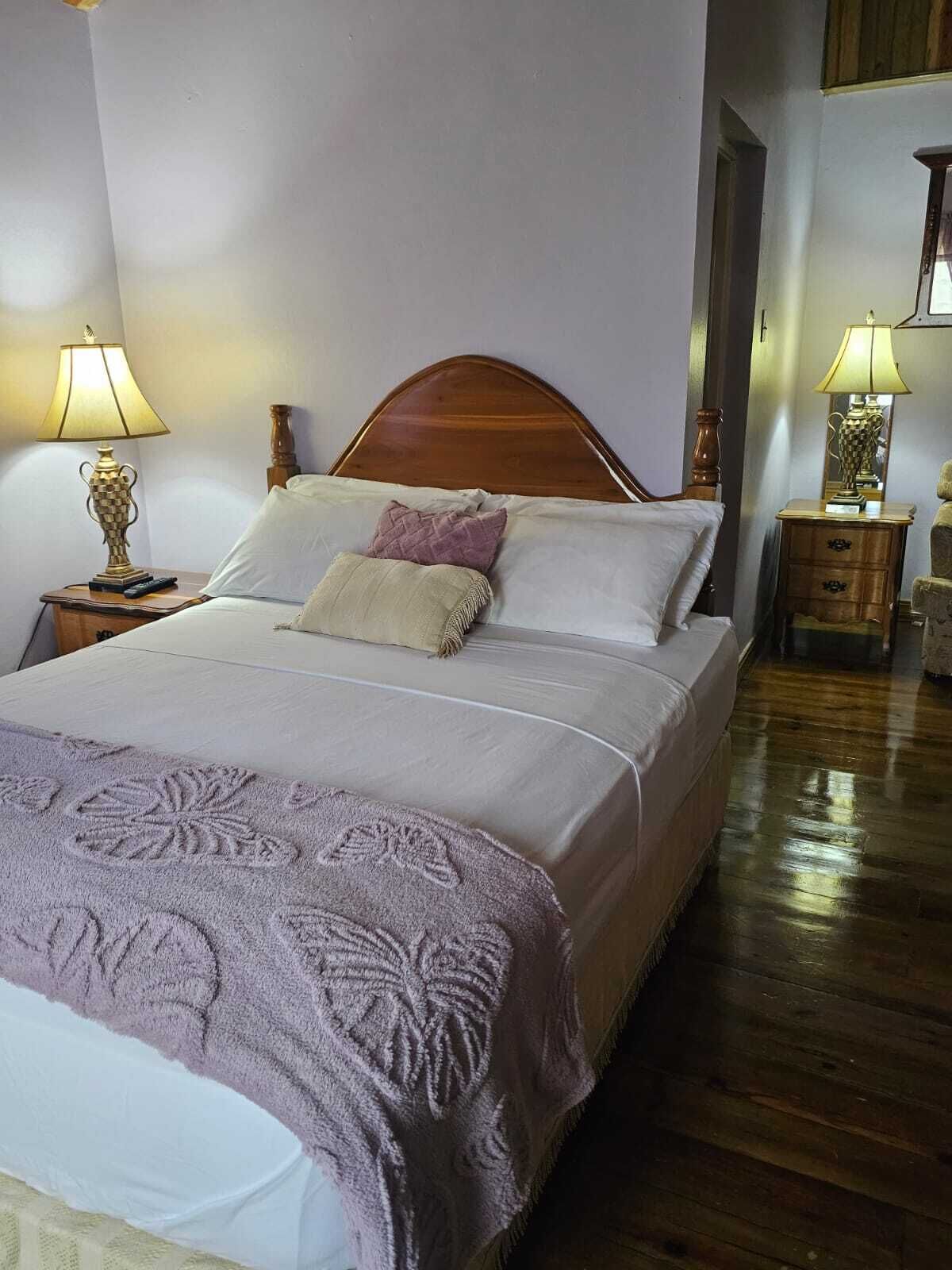 Una cama bien hecha con cabecera de madera, dos mesitas de noche con lámparas y suelo de madera.