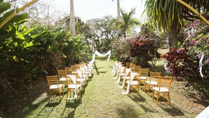 Un salón de bodas al aire libre con filas de sillas de madera y un arco blanco.