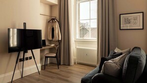 A room with a large window, a black TV on a tripod stand, a chair, a sofa, and a framed picture on the wall.