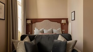 A hotel room with a dark-colored sofa, a headboard with a decorative panel, two wall-mounted lamps, and a framed picture on the wall.