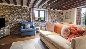 A cozy living room with a stone wall, a blue armchair, a beige sofa, and a television on a wooden cabinet.