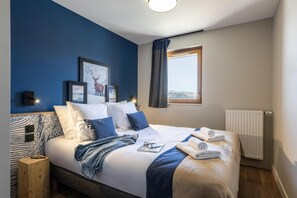 A neatly made bed with white linens, blue pillows, and a blue throw blanket. There are framed pictures on the wall, a window with curtains, and a wooden nightstand with a lamp. - Wifi, ski-in/ski-out & foot of the slopes, swimming pool, balcony, ski locker, 31-34m², Risoul (Risoul)