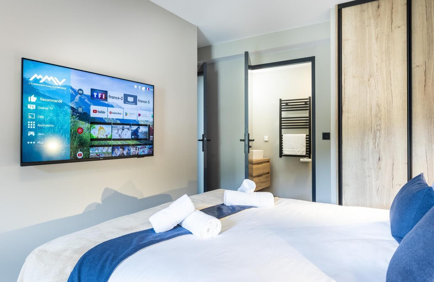 A modern hotel room with a large TV on the wall displaying various channels and apps, a neatly made bed with towels, a wooden nightstand, and a wooden door leading to another room.