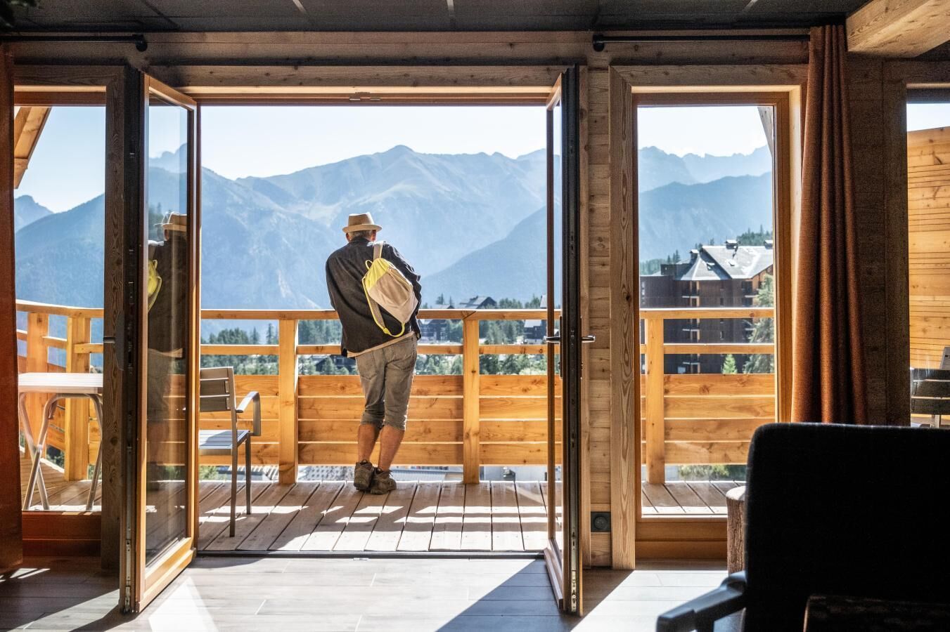 A wooden balcony with a view of mountains, a person standing, and a chair.
