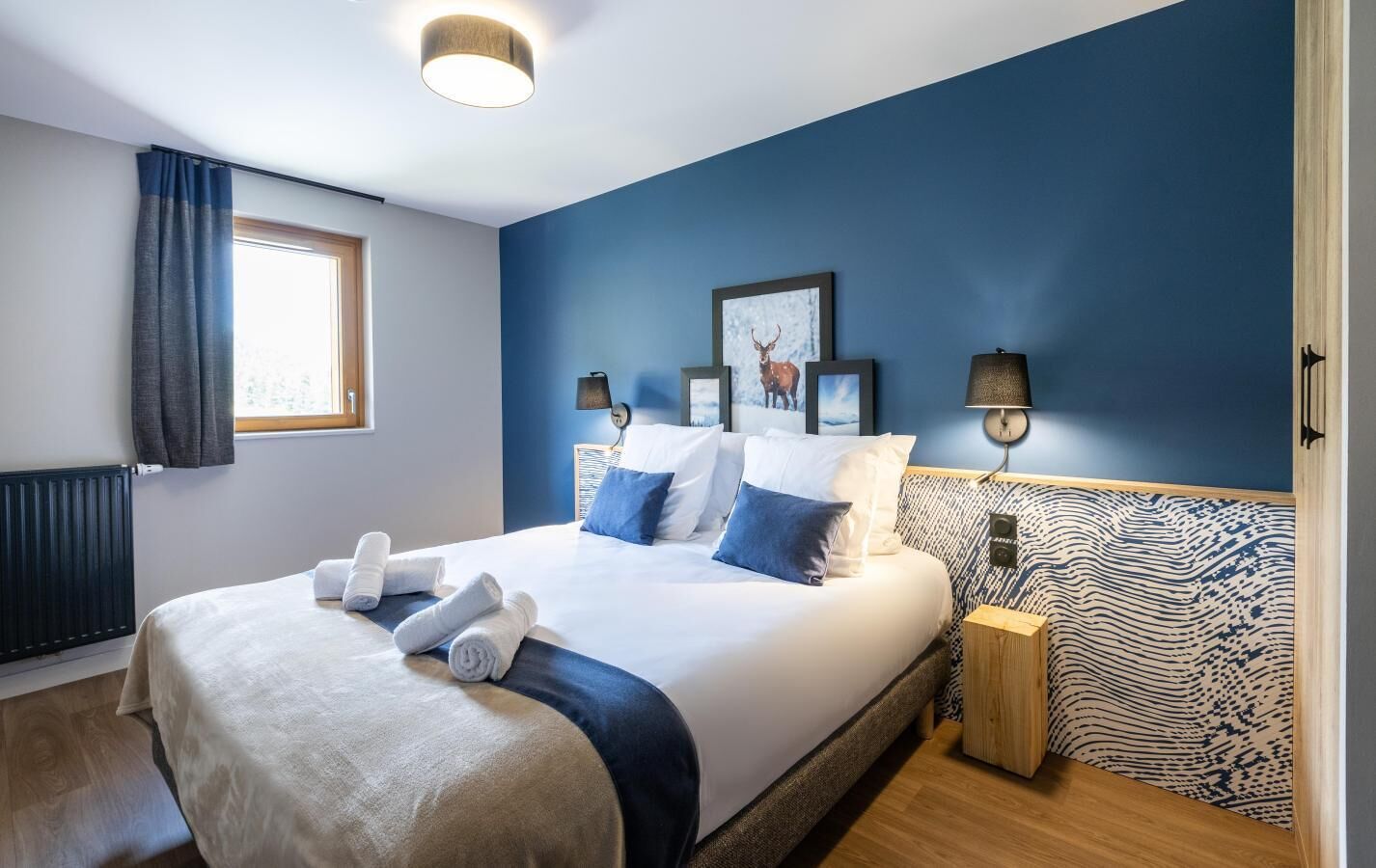 A modern hotel room with a large bed, two bedside tables, a window with curtains, and wall-mounted artwork.