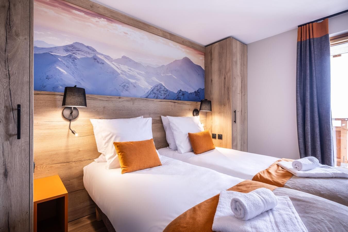 Een hotelkamer met twee bedden, een houten hoofdbord, een schilderij met uitzicht op de bergen en een balkon met gordijnen.