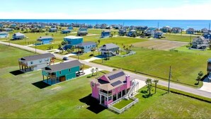 An aerial view of a residential area with houses, a large open grassy field, and a body of water in the background. - The Pink House - beautiful beach stay! (Galveston)