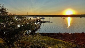 A sunset view over a calm harbor with boats, a wooden pier, and surrounding vegetation.