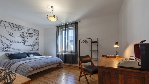 Ein Schlafzimmer mit einem Bett, einem Schreibtisch mit Schreibmaschine, einem Stuhl, einer Lampe und einem Wandbild.