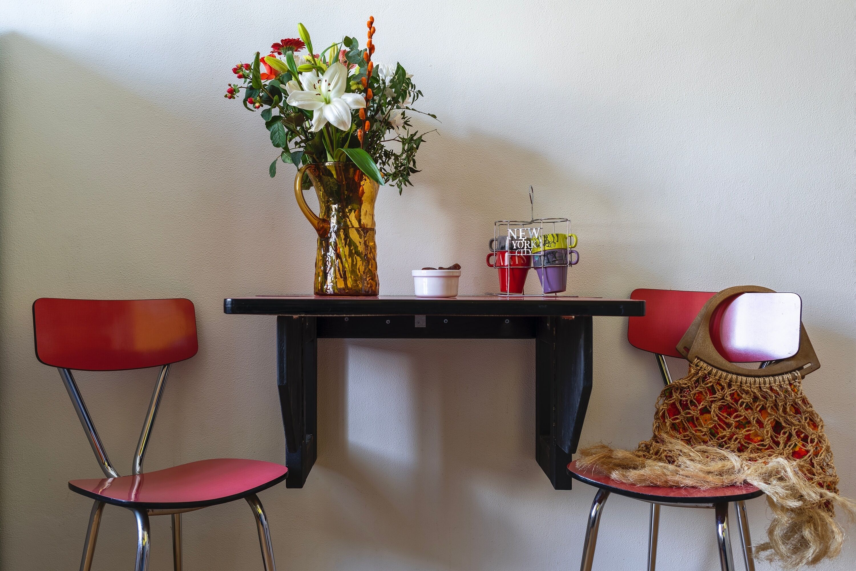 A table with a vase of flowers, a coffee maker, and a mug. Two red chairs are positioned around the table.