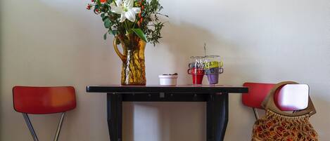 A table with a vase of flowers, a coffee maker, and a mug. Two red chairs are positioned around the table.