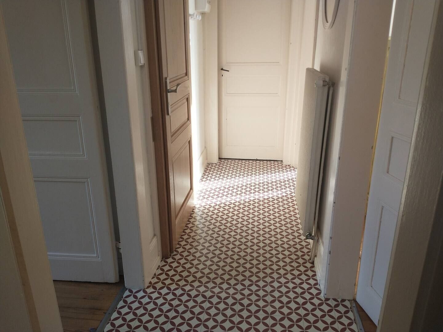 A narrow hallway with patterned floor tiles, wooden doors, and a radiator on the right wall.