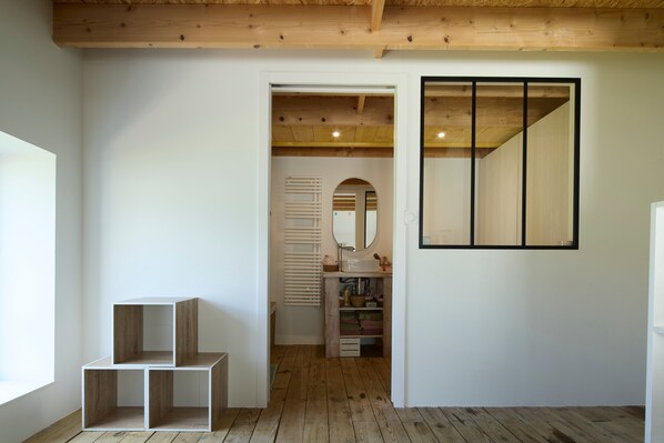 Una habitación con techo de madera, pared blanca y un espejo grande. Hay dos estantes de madera y una puerta que da a otra habitación.