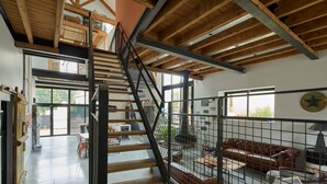 Een ruime loft met een hoge plafond, zichtbare houten balken, een trap met een metalen leuning en een leren bank in de woonkamer.