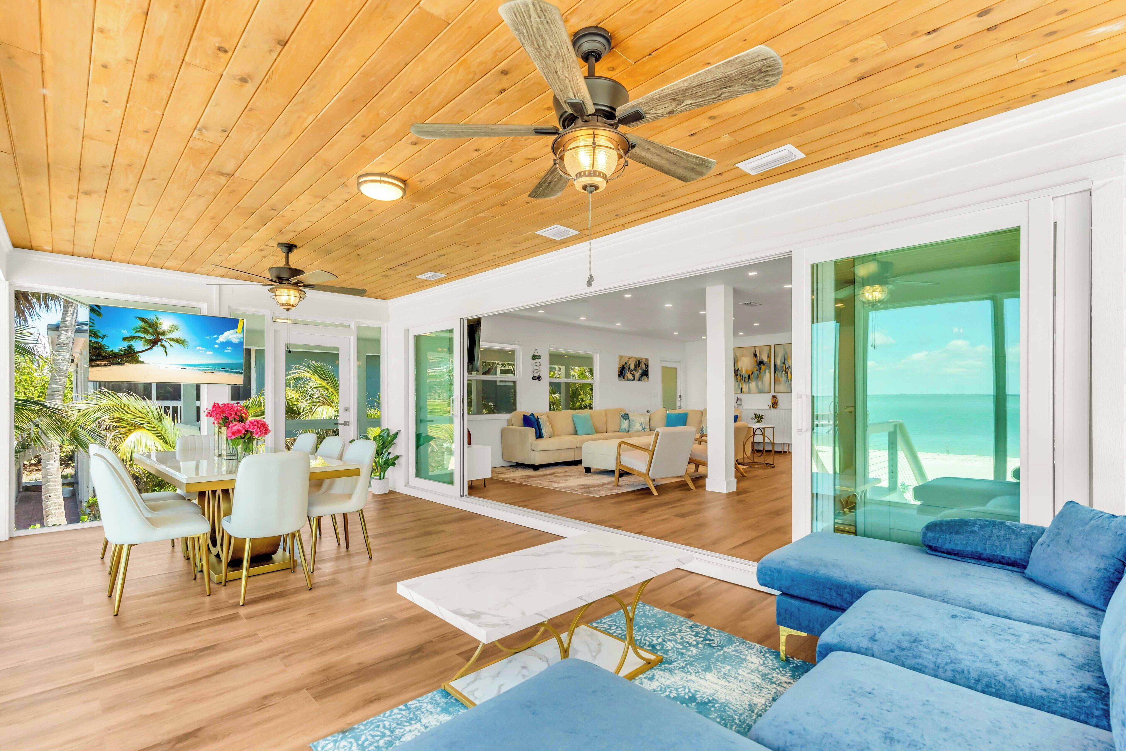 A spacious living area with a wooden ceiling, a dining table with chairs, a blue sofa, and a view of the ocean through large glass doors.
