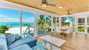 A spacious living area with a large window overlooking a beach, a ceiling fan, and a dining area with a table and chairs.
