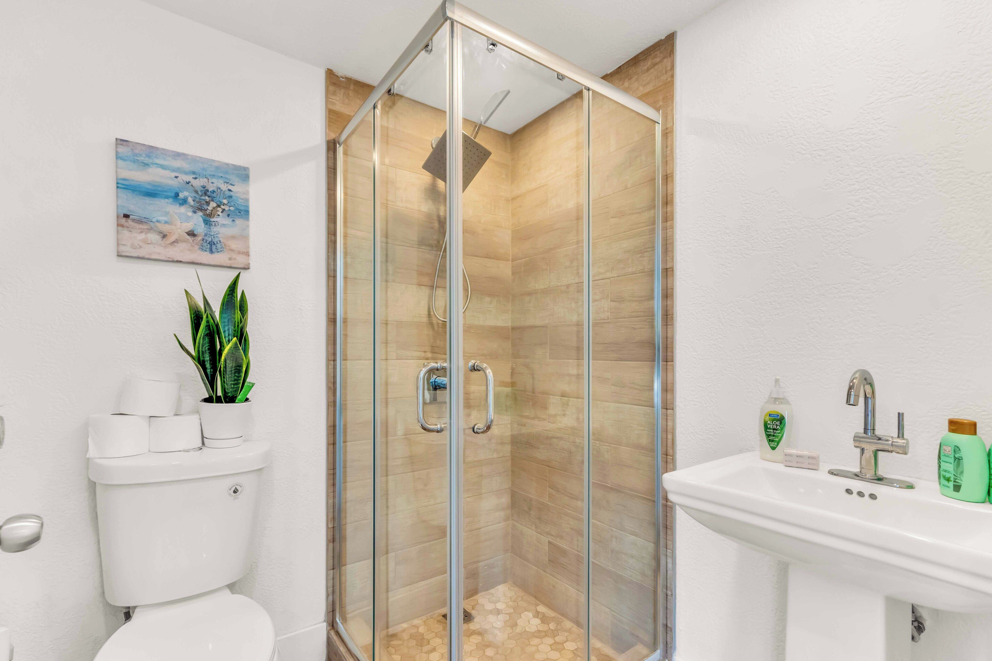 A modern bathroom with a glass-enclosed shower, a toilet, a sink with toiletries, and a painting on the wall.