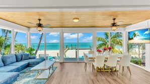 A spacious room with a large window overlooking a beach, a dining area with white chairs, and a blue sofa.