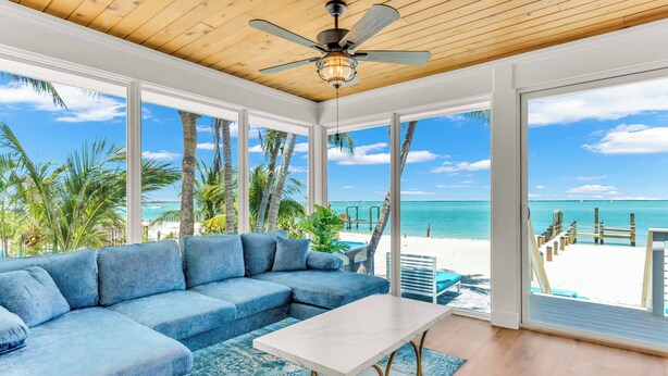 A room with a large window overlooking a beach, a blue sofa, and a ceiling fan.