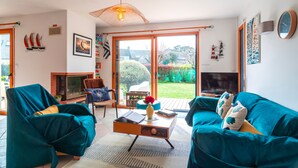 A living room with teal sofas, a coffee table, and a fireplace. There is a large window with a view of a garden. - Ty Kéréré - Maison 3★ - Calme, Grand Jardin Arboré, 500m plage (Locmariaquer)