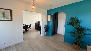 Een modern interieur met een blauwgroene accentmuur, een eethoek met een tafel en stoelen, een potplant en een plafondlamp.