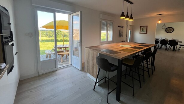 Een moderne keuken met een groot kookeiland, schuiframen naar een terras en een eethoek met een tafel en stoelen.
