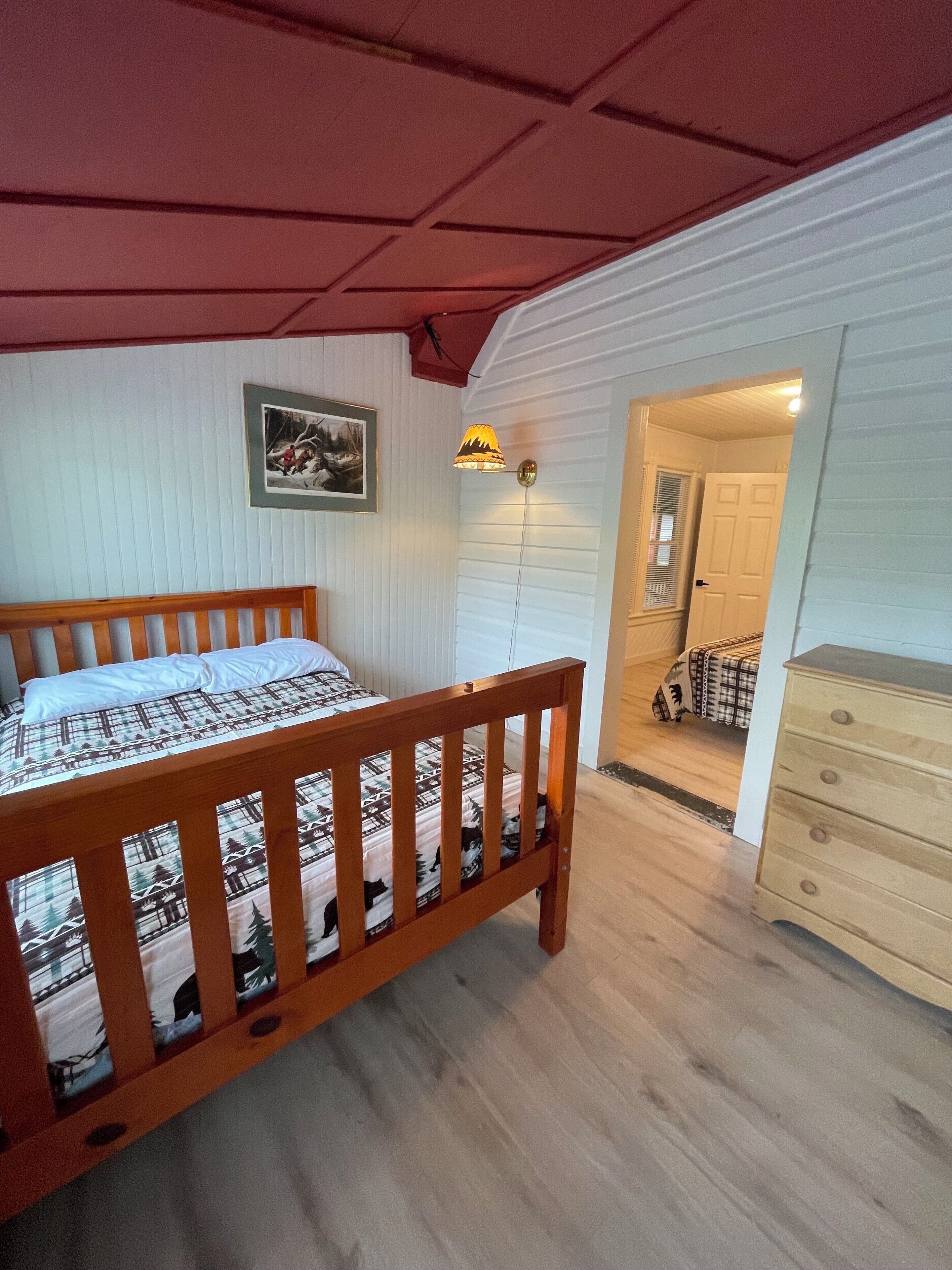 A bedroom with a wooden bed, a wall-mounted lamp, a door leading to another room, and a dresser.