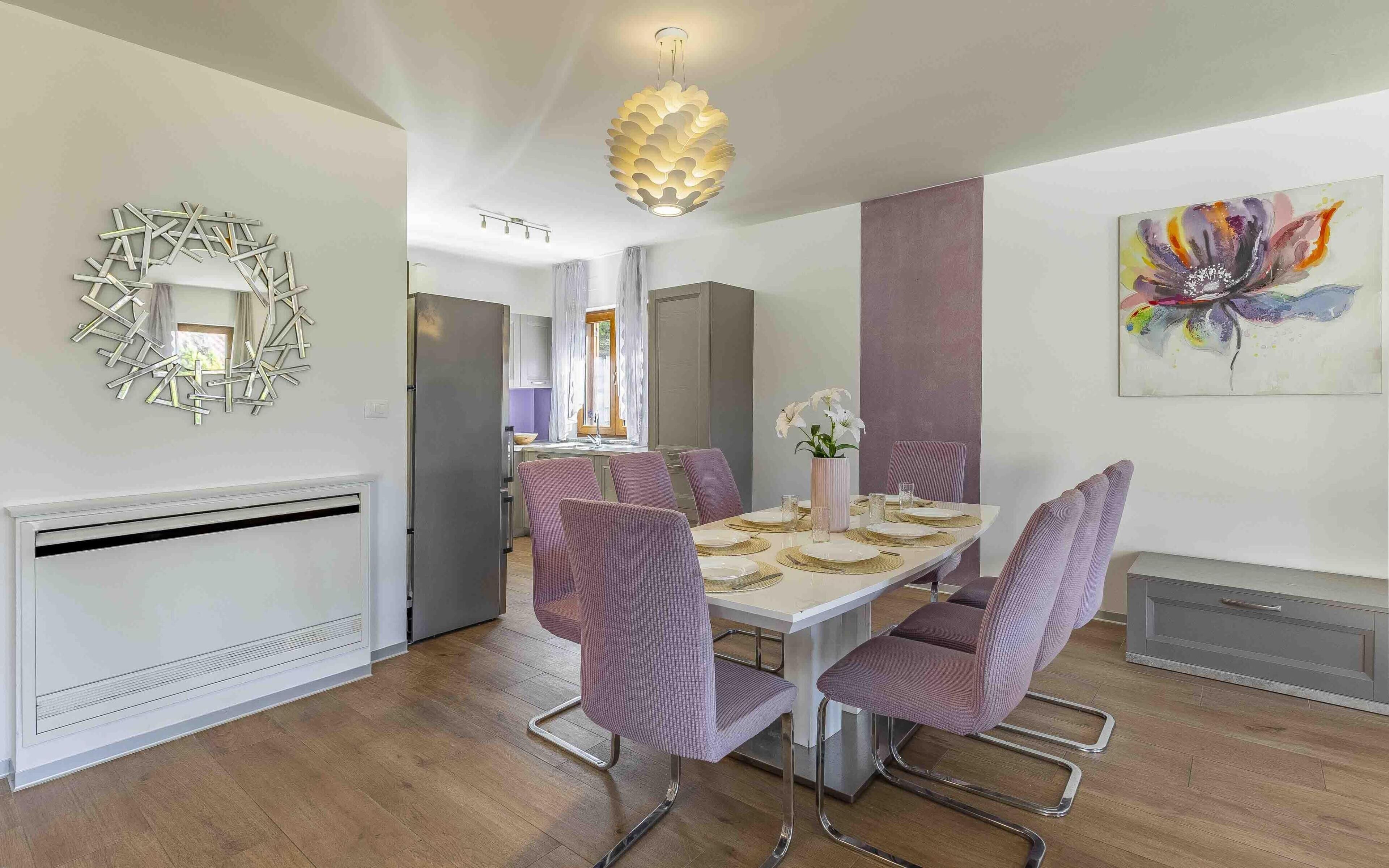 A modern dining area with a white table set for four, surrounded by purple upholstered chairs. A decorative light fixture hangs from the ceiling, and a colorful abstract painting is on the wall.