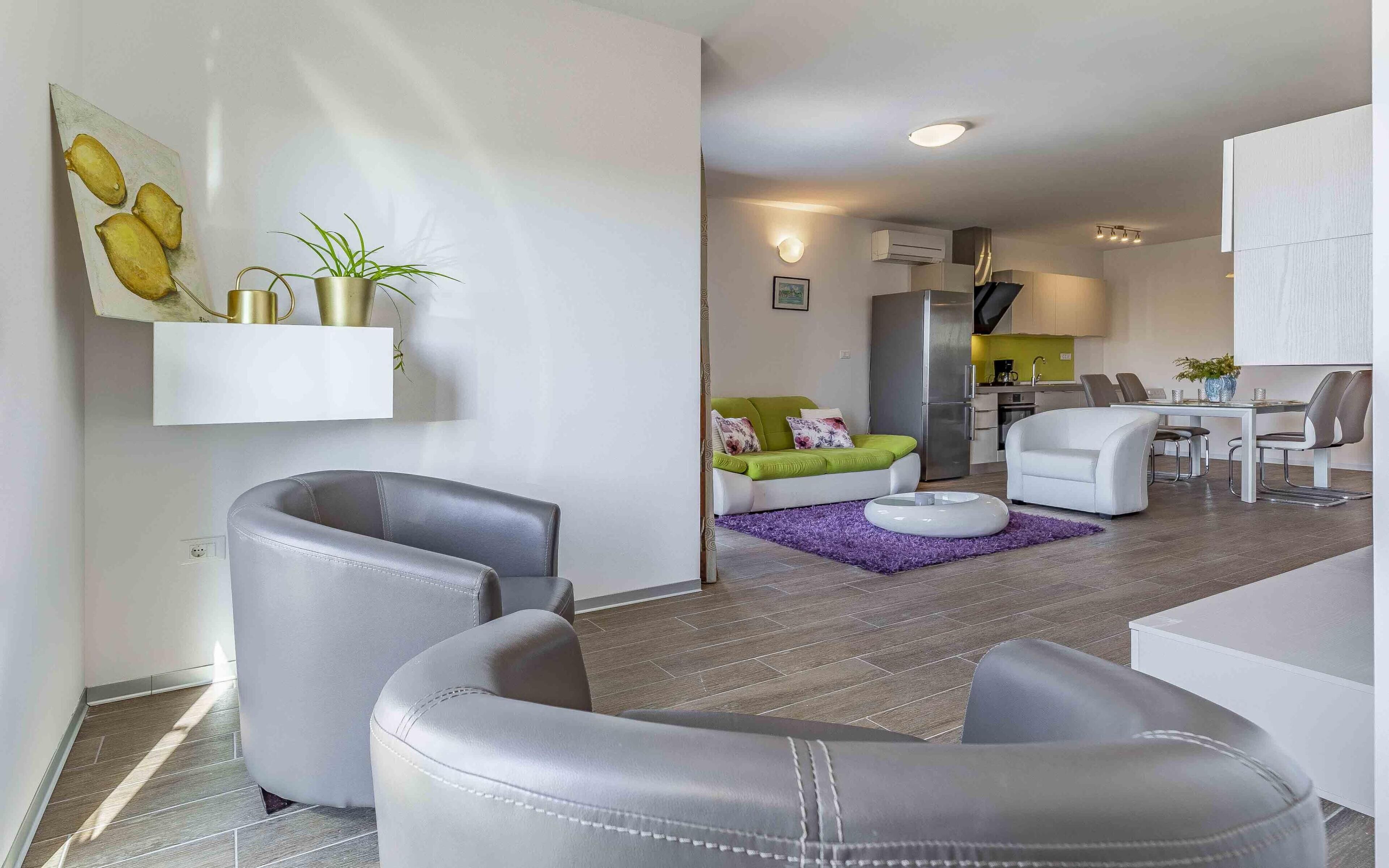 A modern living room with grey leather sofas, a dining area with white chairs, a kitchen with a stainless steel refrigerator, and a small dining table.