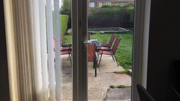 A sliding glass door leading to a patio with a table and chairs, a hedge, and a house in the background. - House-sleeps 5-parking-bbq-garden-patio (Chesterfield)