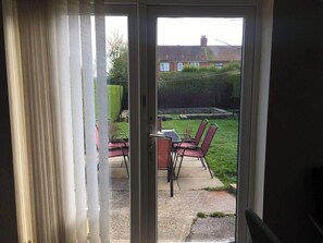 A sliding glass door leading to a patio with a table and chairs, a hedge, and a house in the background. - House-sleeps 5-parking-bbq-garden-patio (Chesterfield)