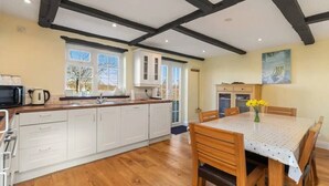 A kitchen with wooden beams, white cabinets, a wooden dining table with chairs, and a window with a view. - Meadow Sweet, Meadowsweet Cottage Jacuzzi Edition (Saundersfoot)
