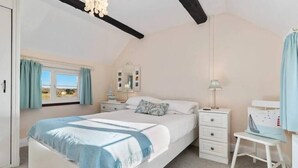 A bedroom with a bed, a nightstand, a dresser, a desk with a chair, a window with curtains, and a chandelier. - Meadow Sweet, Meadowsweet Cottage Jacuzzi Edition (Saundersfoot)