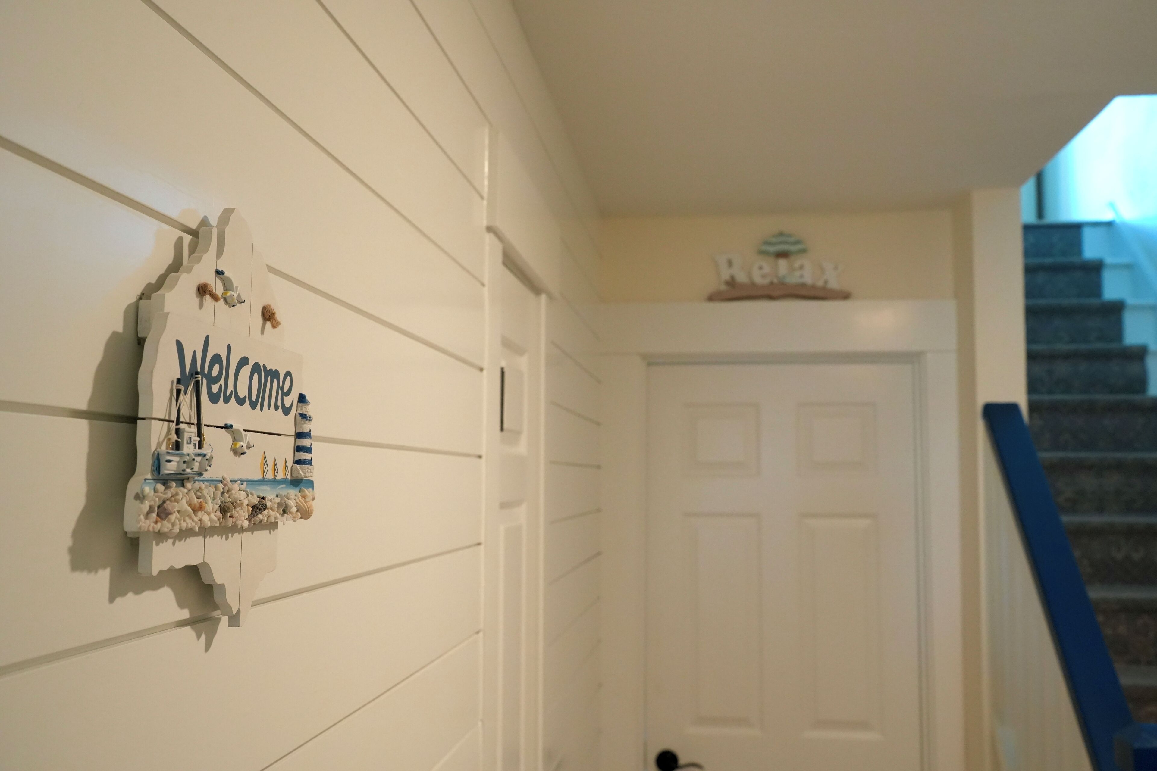 A white wall with a welcome sign featuring a lighthouse and ships, a white door, and a staircase with a blue handrail.