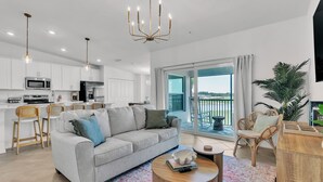 A modern living room with a grey sofa, wooden coffee table, and wicker chairs. A kitchen with white cabinets and stainless steel appliances is visible in the background. - 2 bed 2 bath GOLF condo in Webbs Reserve (Punta Gorda)