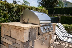 Una barbacoa de pedra, de planta oberta, amb una tapa i quatre fogons, acompanyada d'una cadira relaxant.