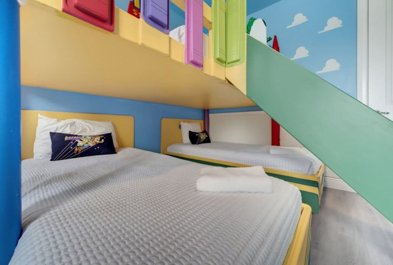 A room with two beds, each with a pillow and a blanket. There is a staircase with colorful steps leading to a door.